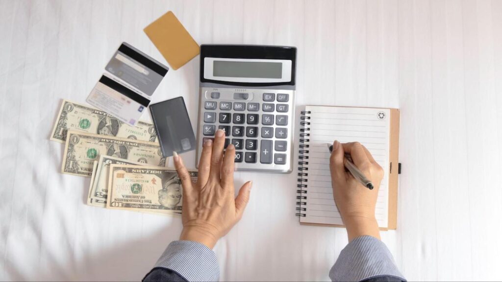 A person types on a calculator and writes in a notebook while multiple credit cards and cash are scattered nearby.