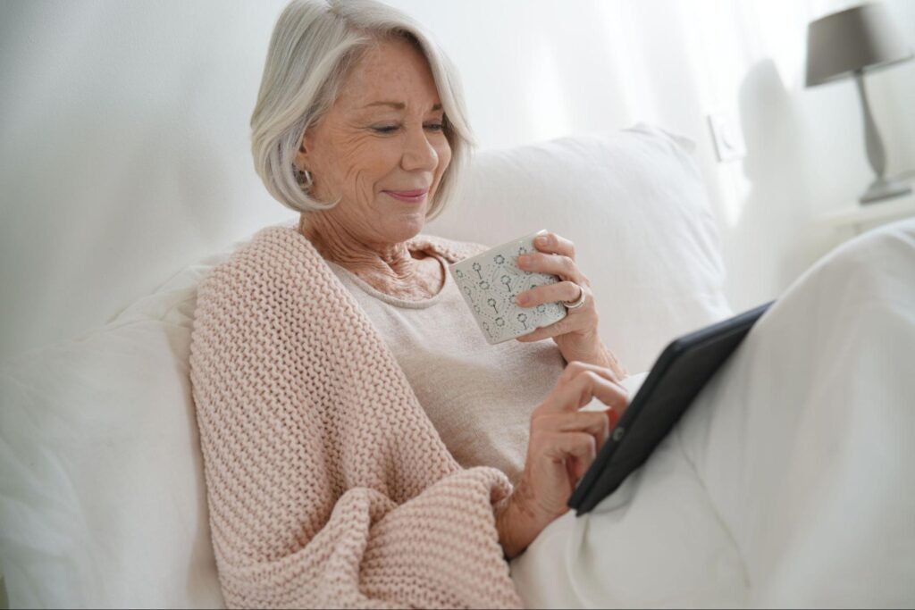 An elderly woman sitting on her couch and managing investments on her tablet device.