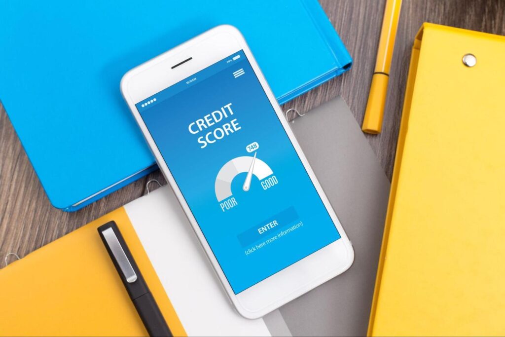 A smartphone lying on top of blue and orange binders with a blue screen that has a credit score meter with the needle leaning towards “Good”.