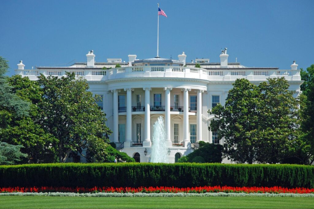 The White House standing behind a large lawn and green trees on a sunny day.