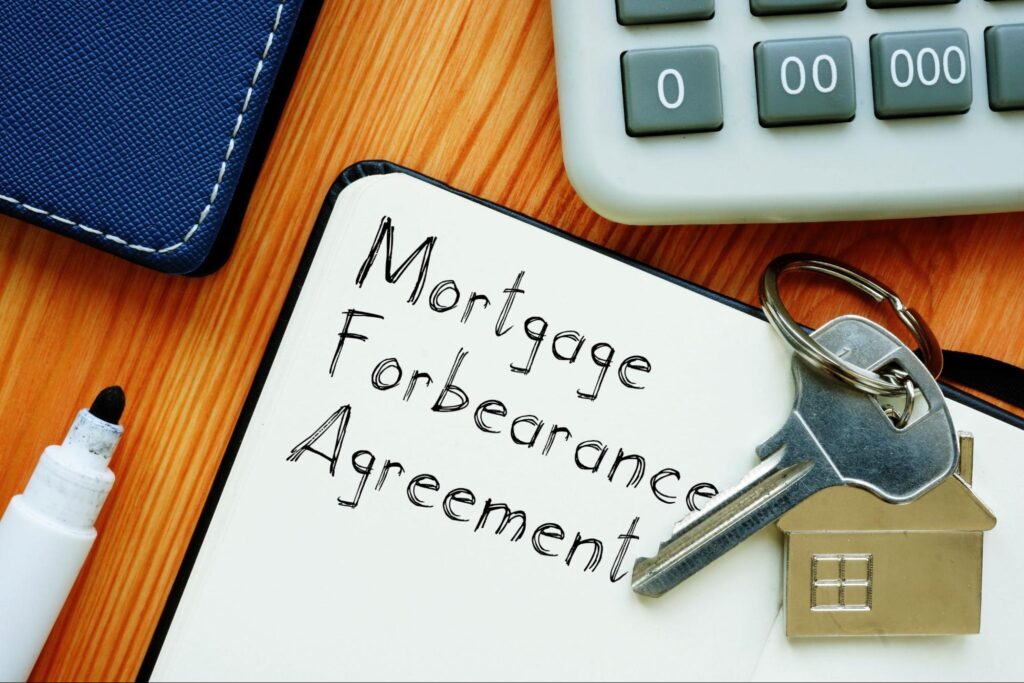 Close-up of a desk with keys, a white board that says “Mortgage Forbearance Agreement”, a calculator, and a pen.