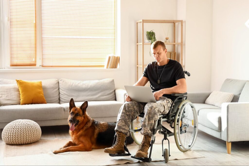 A US military veteran in a wheelchair looks at his open laptop in his living room while his dog sits next to him.