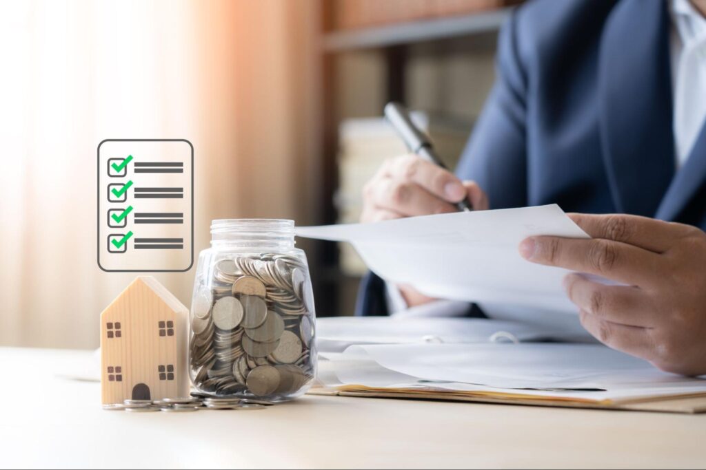 A person signs a document while in the foreground there is a checklist graphic, a small model home, and a jar filled with coins.
