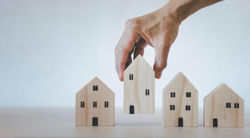 A hand lifting up a wooden model home situated alongside a series of other wooden model homes.