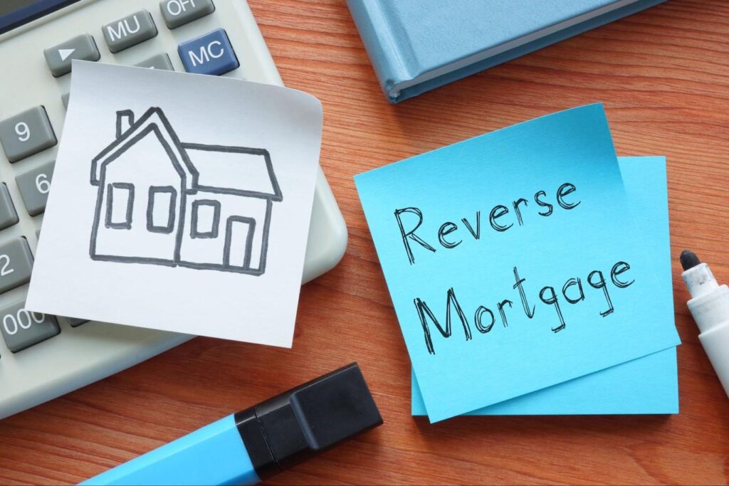 A blue post-it note with “Reverse Mortgage” written on it sits on a wood desk next to a marker, a drawing of a home, and a calculator.