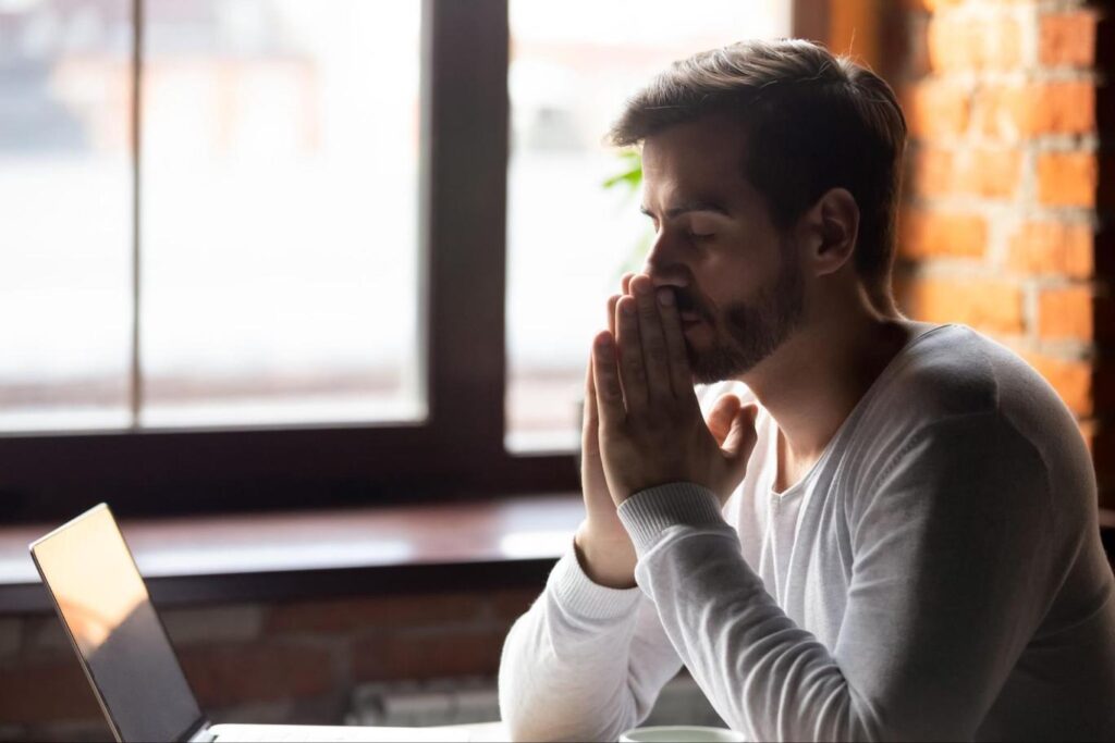 Man sitting in front of a laptop with his hands clasped in front of his face.