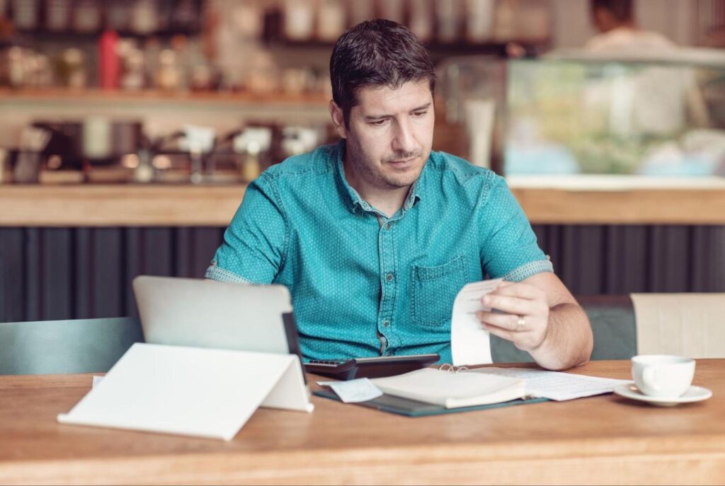 A small business owner in a teal shirt sits at a desk and reviews receipts while filing taxes.