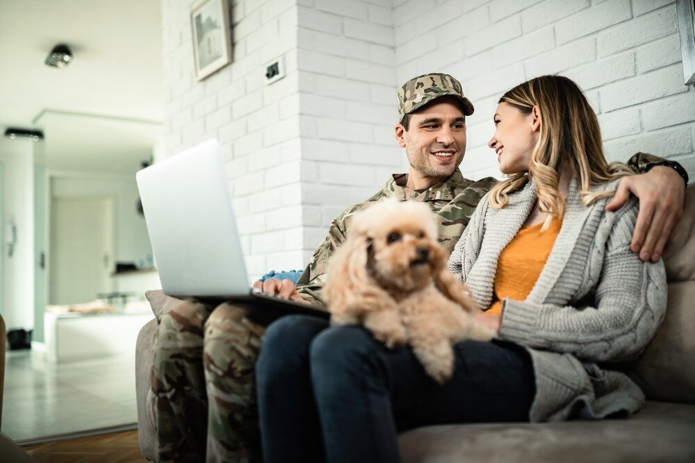 A man dressed in military fatigues sits on a couch with an arm around his wife, an open laptop on his lap, and a small dog sitting on his wife’s lap.