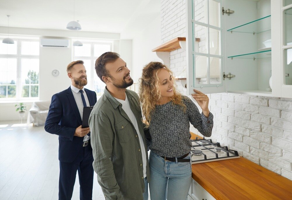 A young white couple tours the kitchen of a bright and clean home with their real estate agent. 