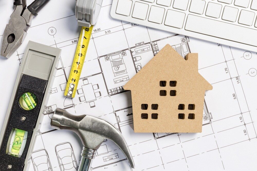 A cutout of a home, a hammer, a leveling tool, a tape measure, pliers, and a keyboard sit on top of a set of blueprints. 