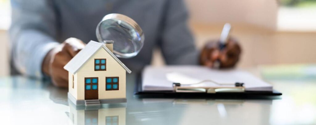 A person writes on a clipboard while holding a magnifying glass up to a cream colored model home.
