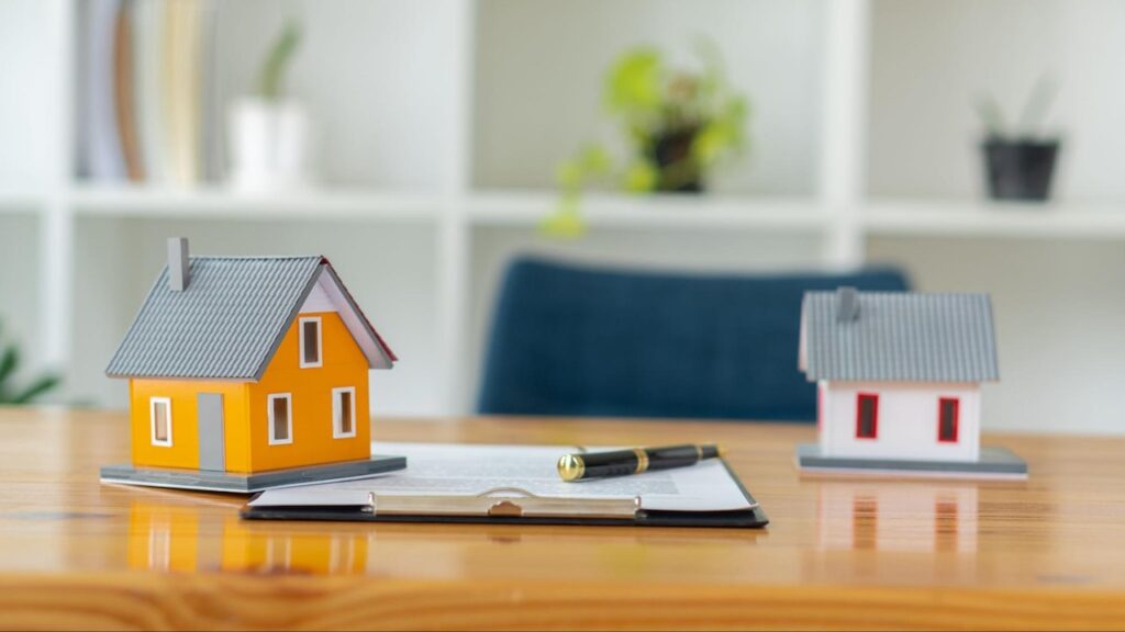 An orange and a white model home sit on a wooden desk alongside a contract on a clipboard.