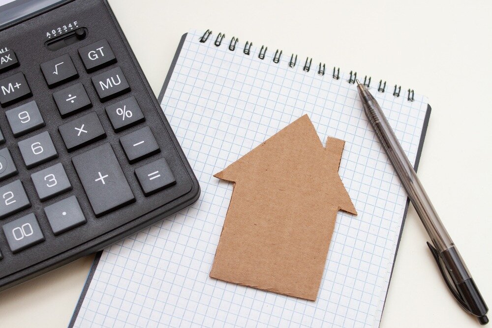 A cardboard cutout of a home sits on top of a notepad and next to a pen and calculator.