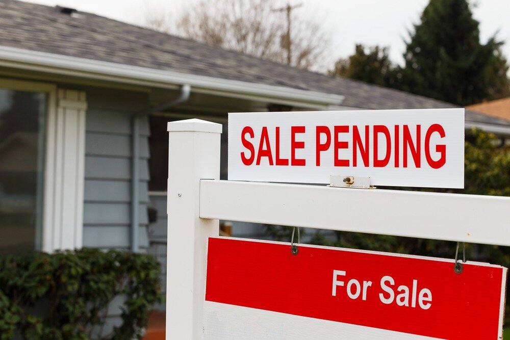 A home’s for sale signpost has a white sign with “SALE PENDING” in red text positioned on top of it.