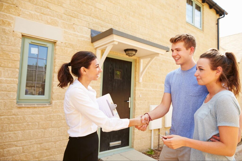 A young happy couple stands outside a tan colored home and shakes hands with a female real estate agent carrying a clipboard under her arm.