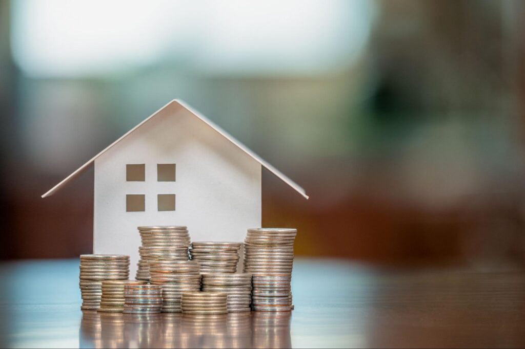 A small white model home positioned behind stacks of coins.