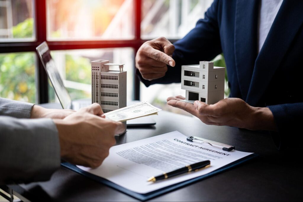 A person in a blue blazer holds a small model home and points to it with their other hand while another person extends their hands over a contract and hands them cash.