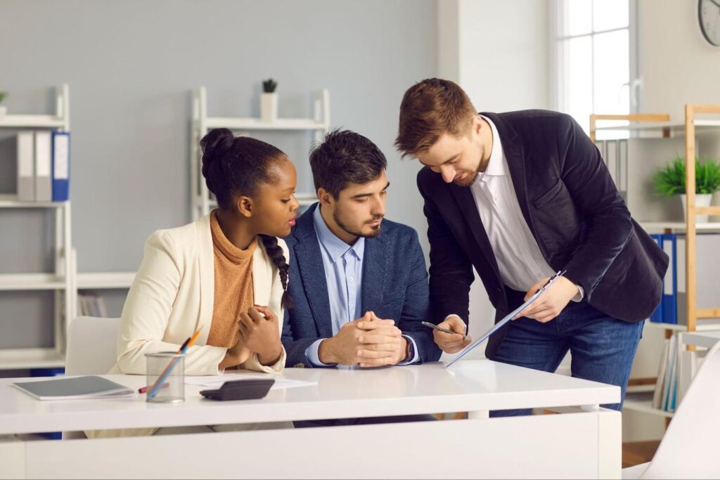 A man in a dark blazer leans over a desk towards a man and woman and points to something on a clipboard.