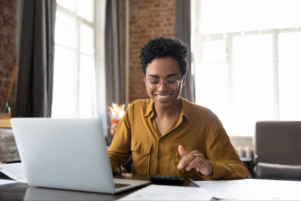 A smiling black woman with short hair sits at a table crowded with papers and simultaneously uses a calculator and her laptop.