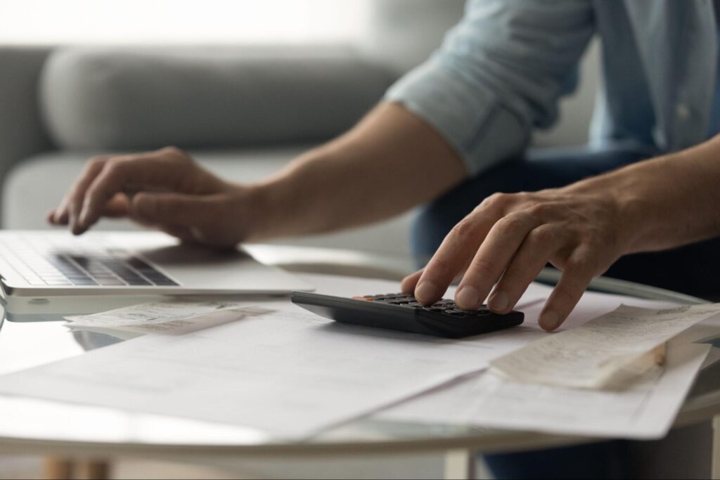 Close up of man’s hands as he types on a keyboard and calculator with papers spread out in front of him on a table.
