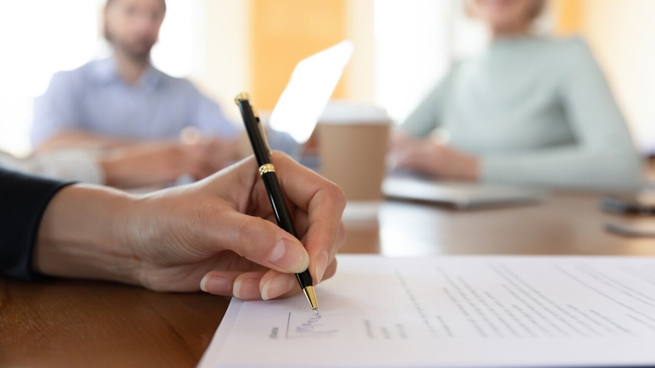 Close up of a woman’s hand signing a document with two people sitting in the blurred background.