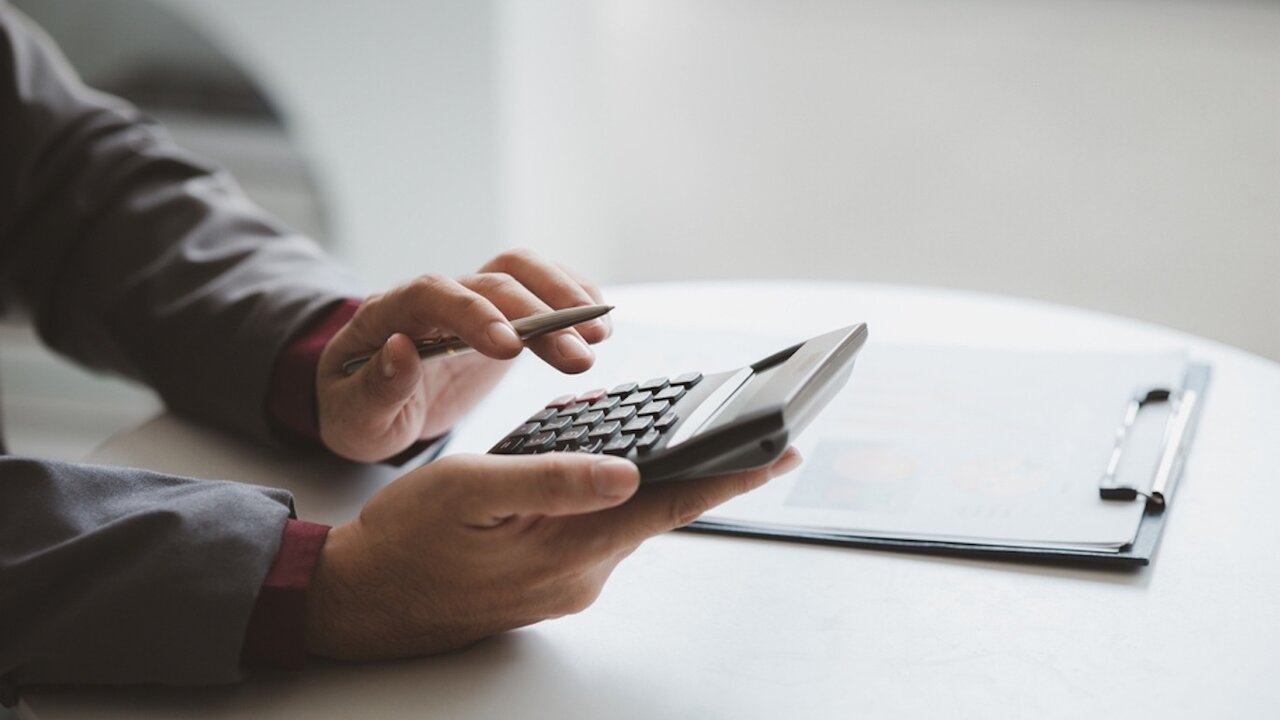The hands of a person holding a calculator over a table that has a clipboard with a document attached to it.