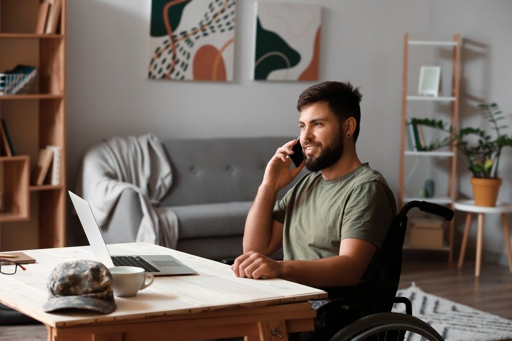 A bearded military veteran in a wheelchair sits at a desk with an open laptop while talking on his cell phone.
