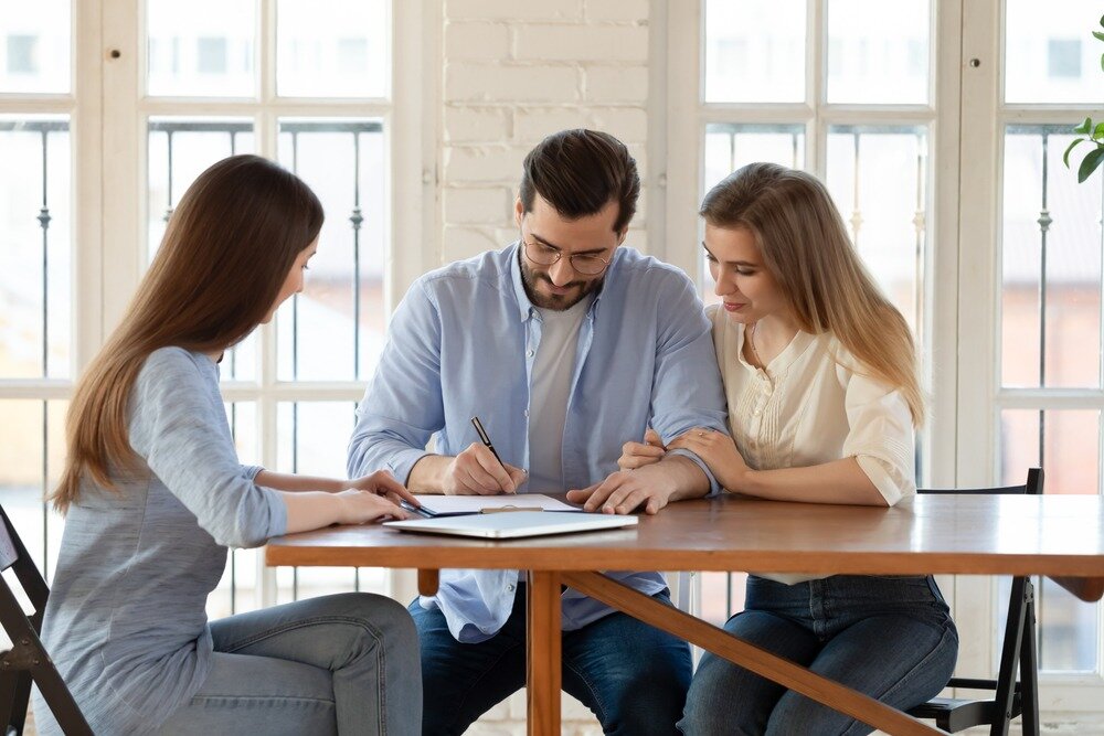A man in a blue collared shirt signs a contract while one woman holds his arm and another woman points to where he should sign. 
