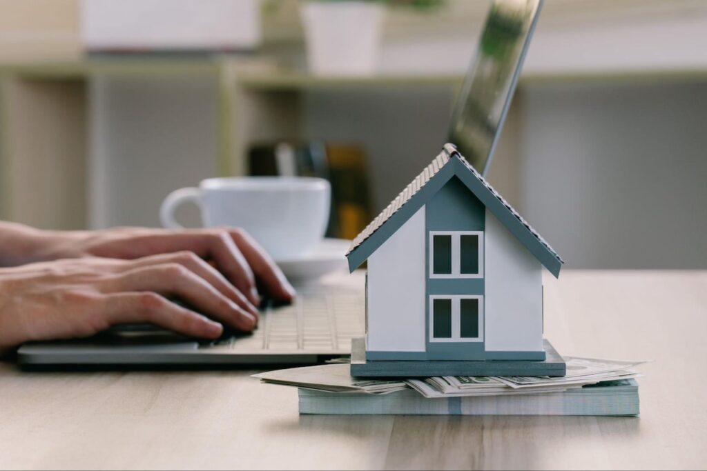 A person types on their laptop next to a small model home sitting on a stack of money.