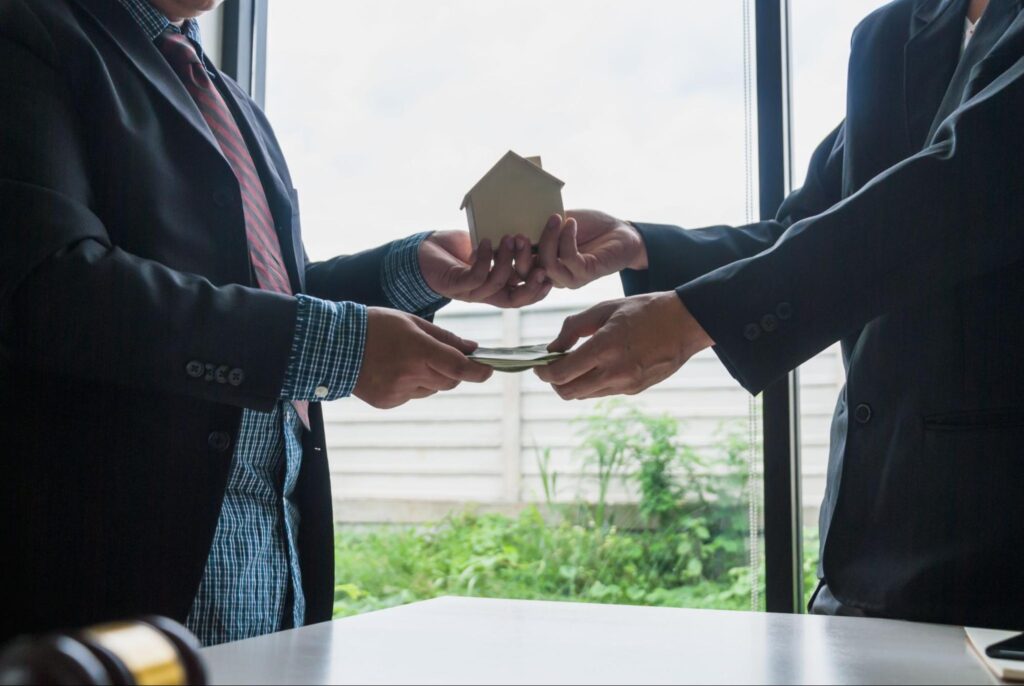 Two people in dark suits stand face to face and exchange a model home with one hand and money with the other hand.