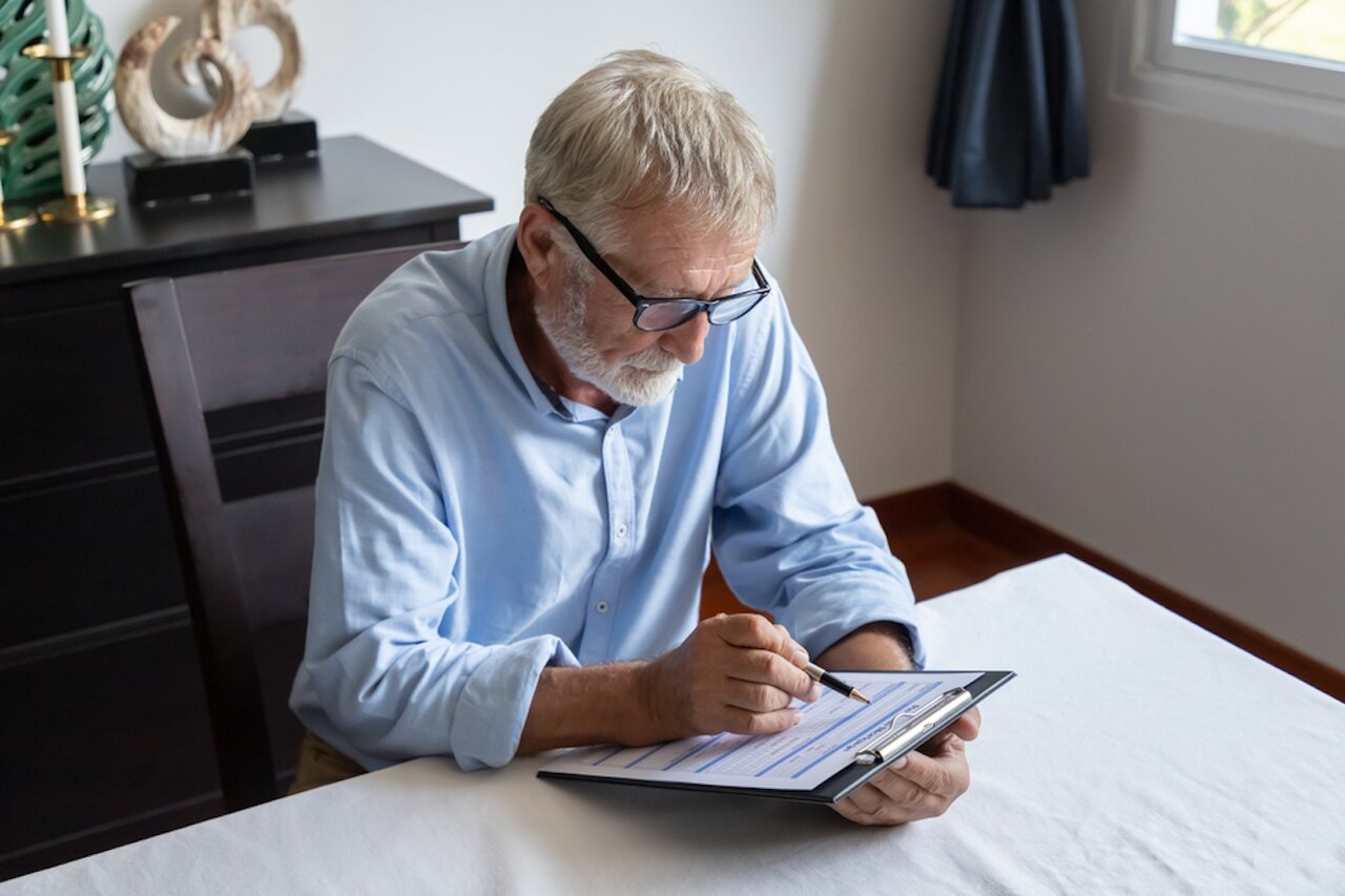 An older man in a light blue collared shirt and glasses sits at a table and reads over a document attached to a clipboard.