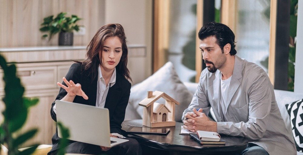 A woman in a suit sits across a small table from a man in a gray blazer and gestures towards her laptop.