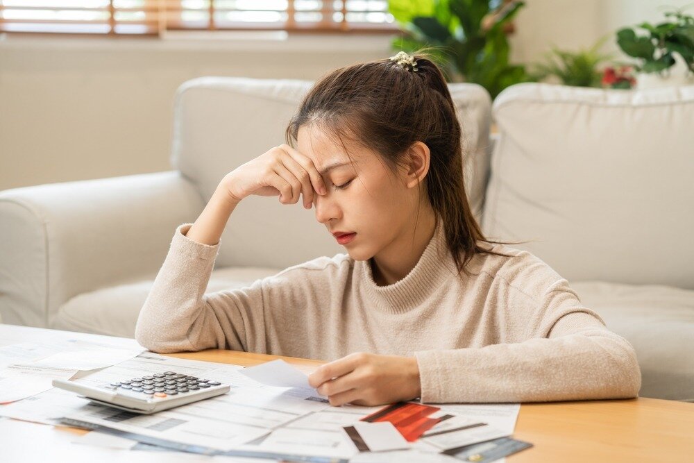 A stressed out Asian woman sits in front of a coffee table cluttered with a calculator, financial documents, and credit cards.