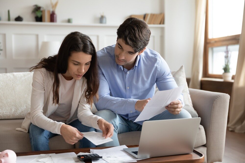 Couple sitting on the couch and managing their finances with a laptop and paperwork spread out in front of them.