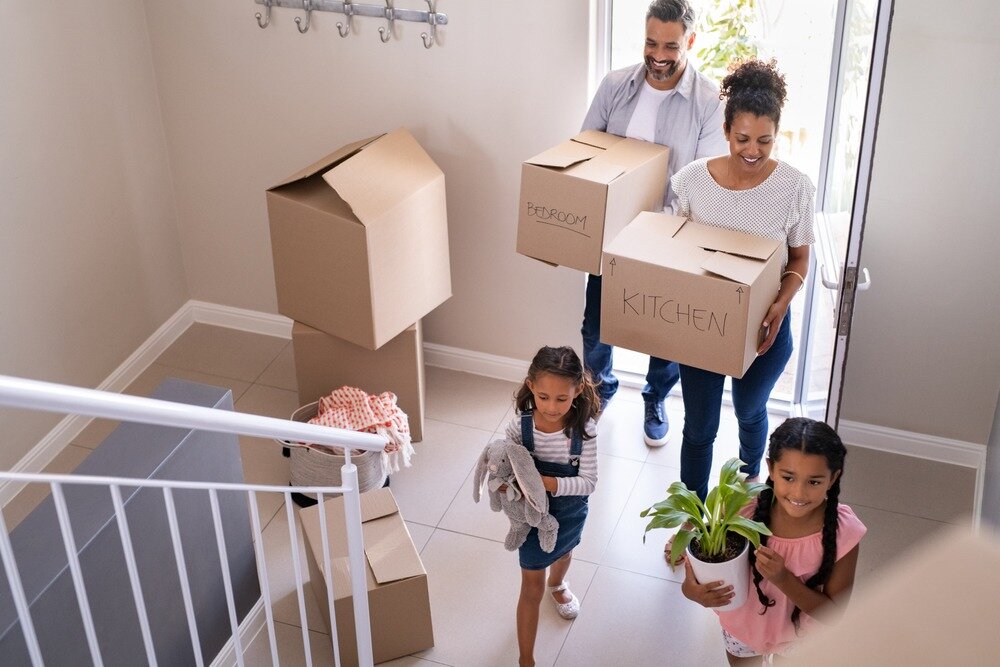A man and a woman carrying cardboard boxes walk into a new home with their two young daughters ahead of them.