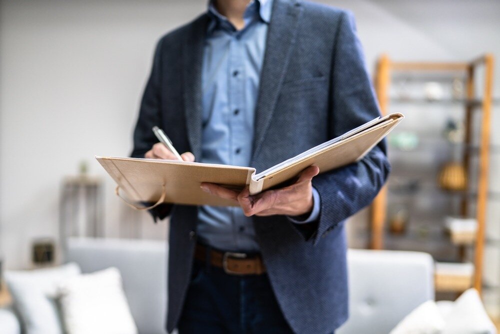 A home appraiser in a blue button up shirt and blazer stands in the room of a home and writes on a clipboard.