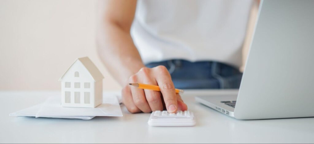 A person at their laptop holding a pencil uses a small white calculator next to a small white model home on a stack of papers.