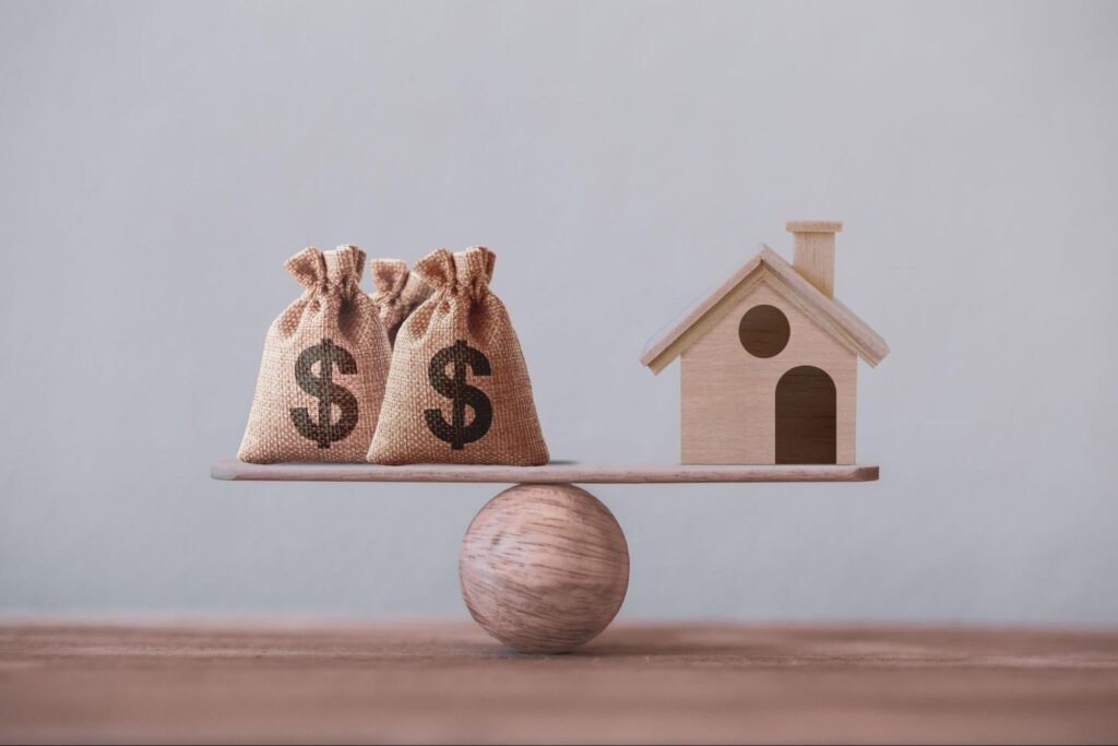 Bags of money and a small model home on opposite sides of a balance scale.