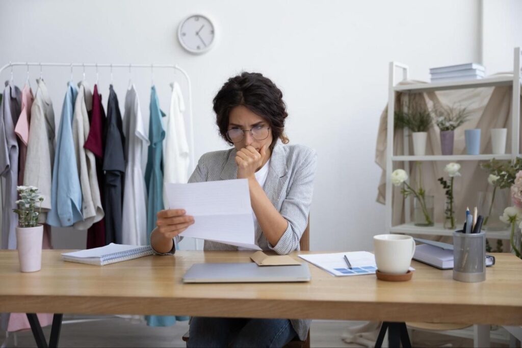 A white woman sits at a desk in her shop and reviews a document with a concerned look on her face.