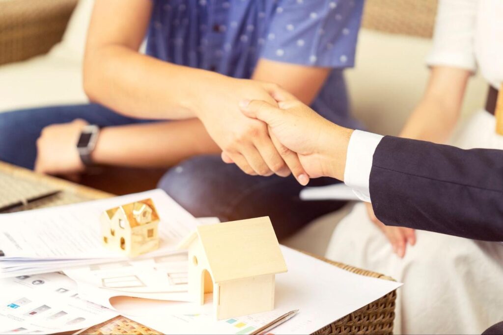 Two people shake hands over a pile of paperwork and two small wooden models of houses.