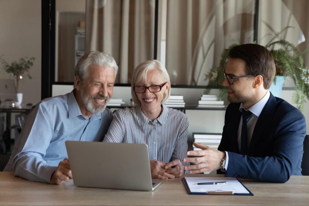 Senior couple sitting in office talking to loan specialist with clipboard and laptop in front of them.