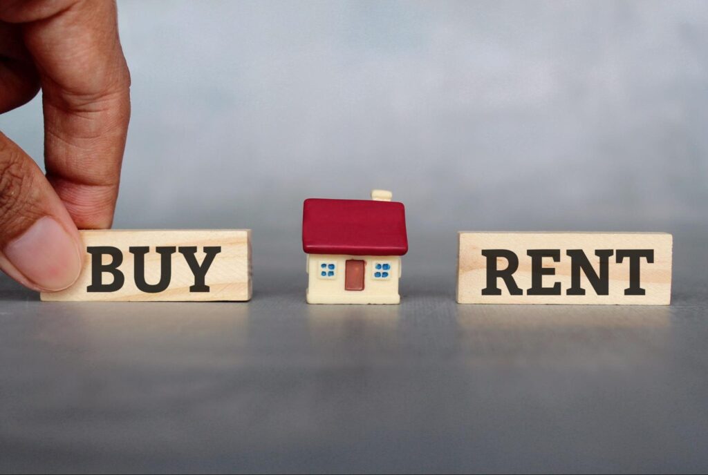 A hand puts down a wooden block that says “Buy” on the opposite side of a model house from a wooden block that says “Rent”.