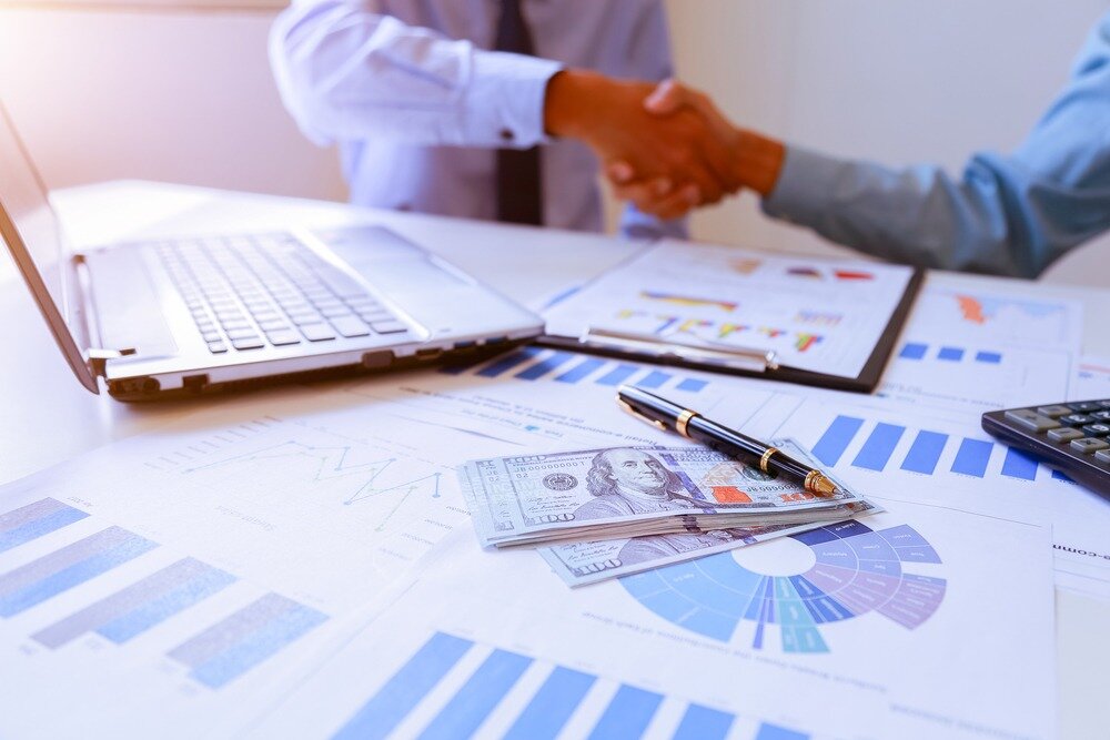 Two people shake hands next to a table upon which is a laptop, several documents with graphics, a stack of money, and a calculator. 