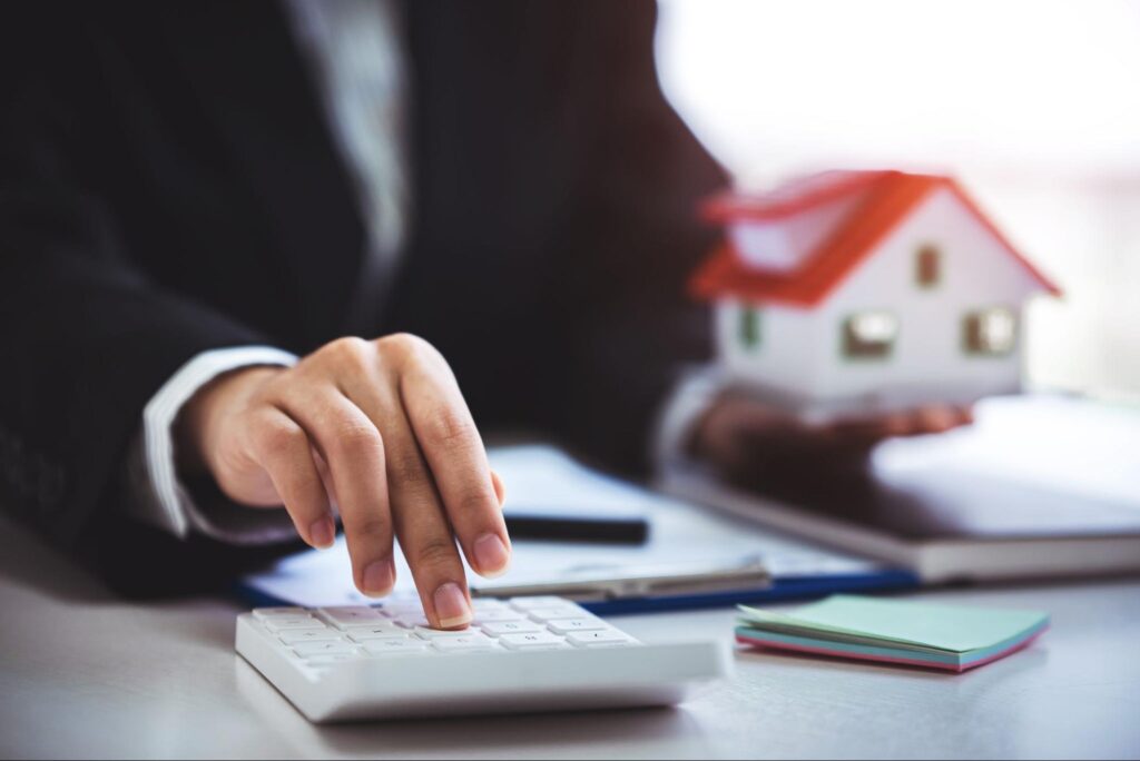 A person in a suit types on a calculator with one hand and holds a model home in the other hand.
