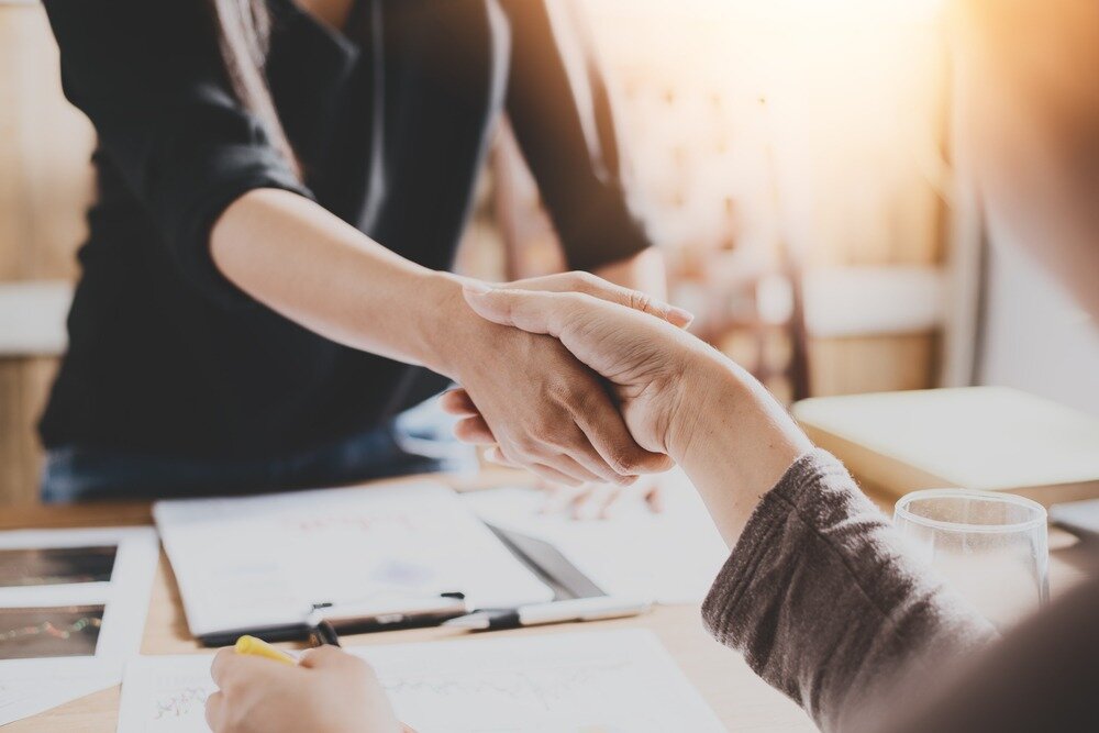 Close up of two women shaking hands making an agreement. The desk in front of them is covered in paperwork.