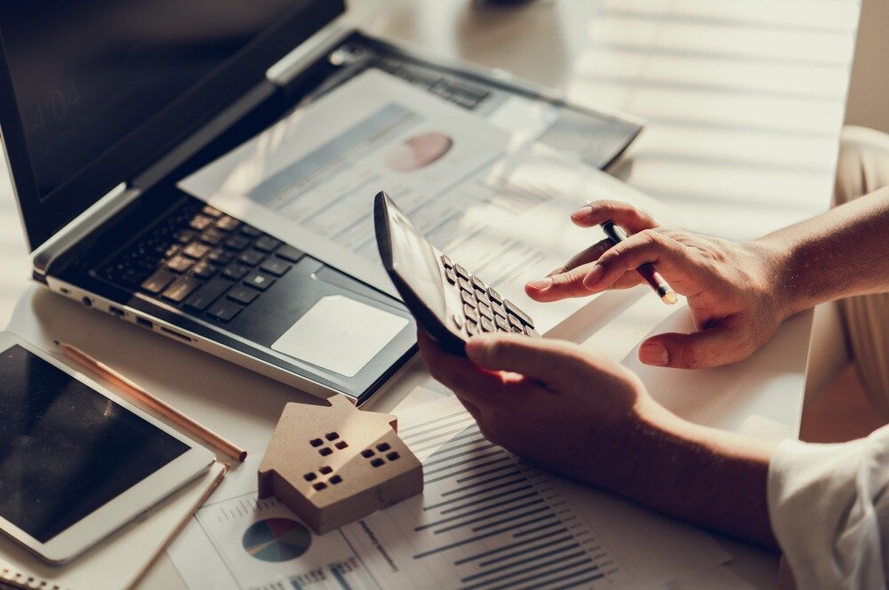 A person types on a calculator over a desk that has an open laptop on it, a small cutout of a house, and several charts and graphs.