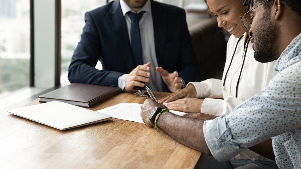 A man with rolled up sleeves signs a document as a woman watches and a man in a navy suit sits on the other side of the table.