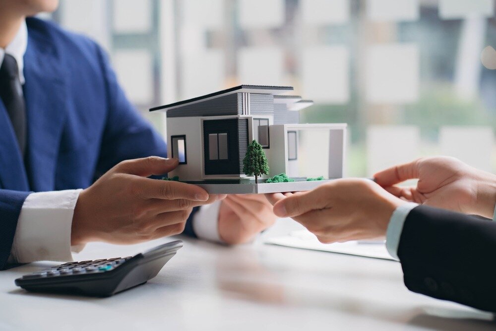 A person in a suit passes a model home over to another person in a suit. 