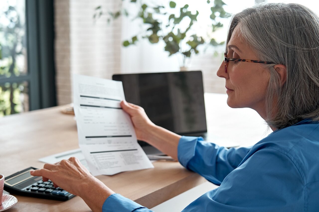 Mature woman using a calculator and holding a paper form in her hand while sitting at a desk.