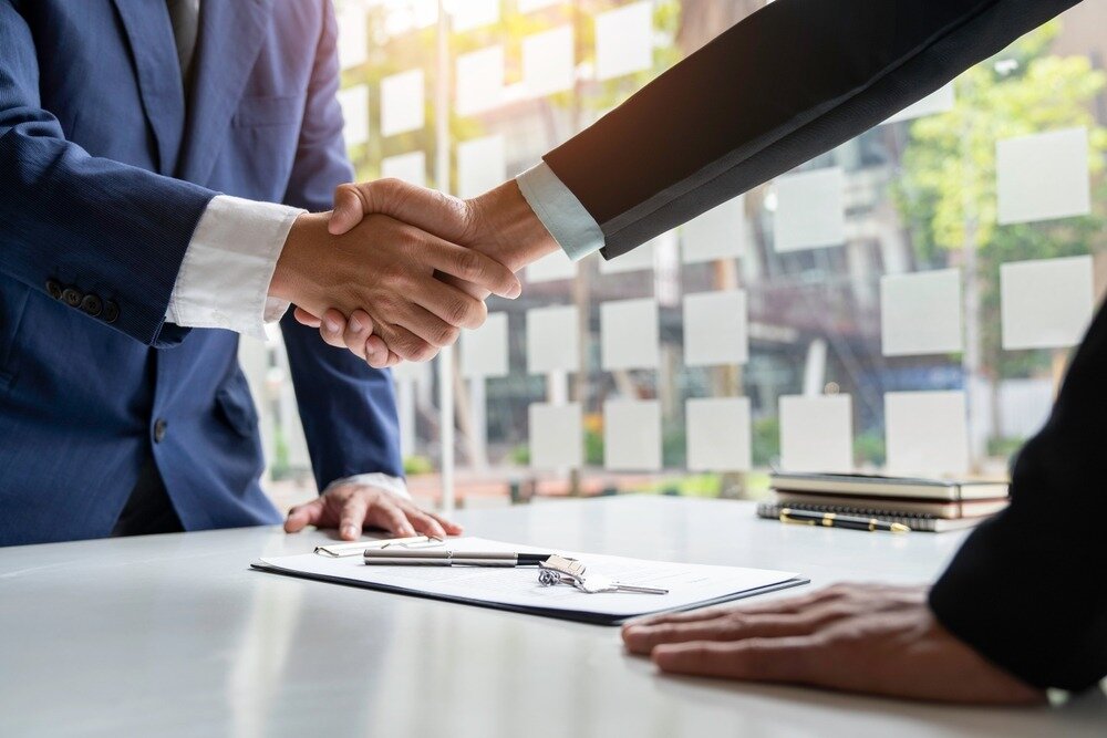 Two people in suits reach across a table and shake hands over a signed contract and house keys. 